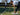 Woman tying shoelaces on tennis court holding a racket, man practicing in the background during tennis training session