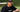 Man wearing black Mantis Snapback Cap and matching black Mantis t-shirt sitting on green bench at tennis court