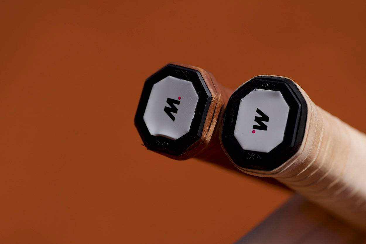 Close-up of two tennis racket handles with replacement grips and M logo end caps on brown background