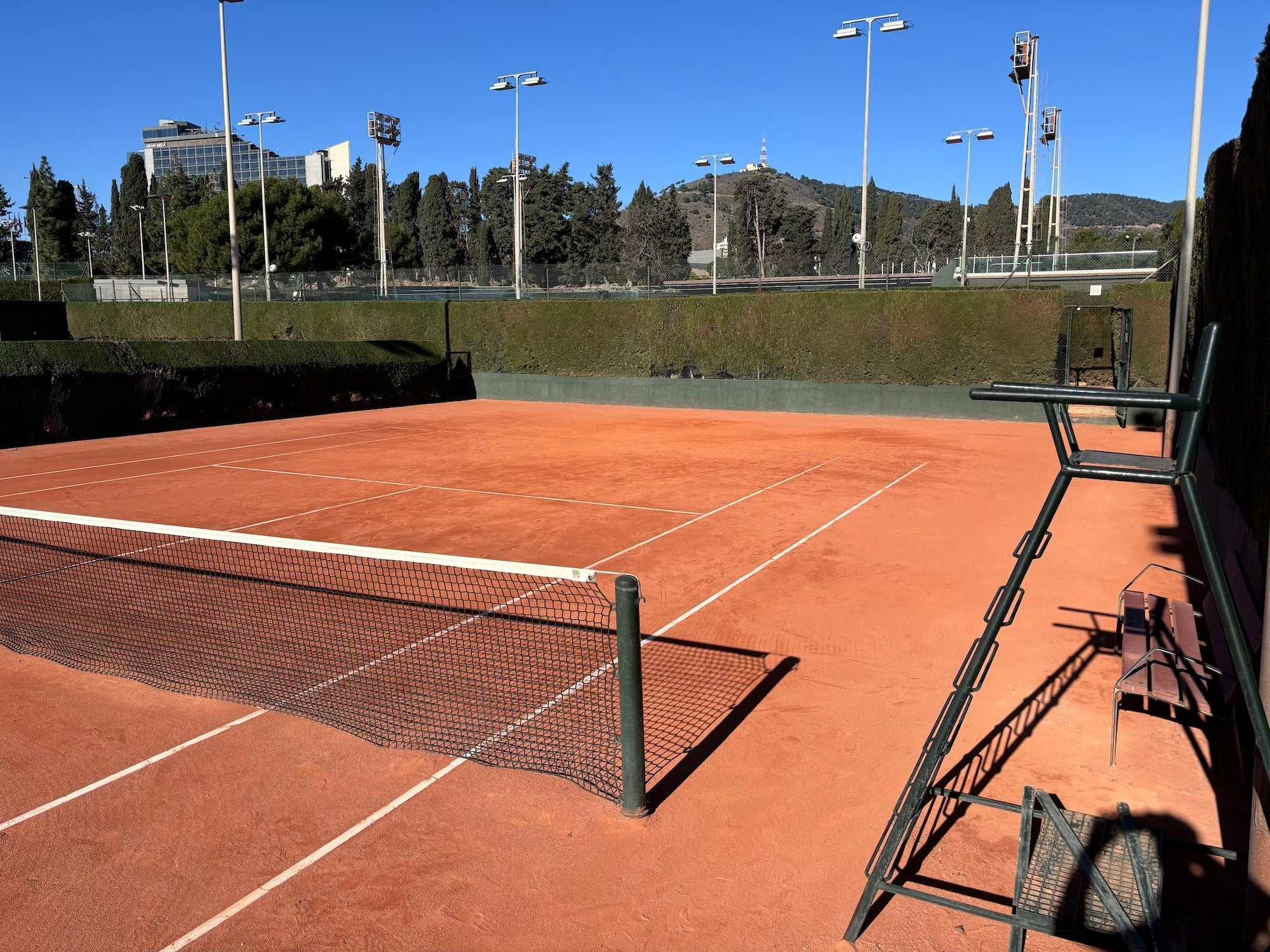 Empty clay tennis court with net and chair umpire stand under clear sky, ideal for tennis lessons for adults