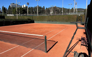 Empty outdoor clay tennis court with standing umpire chair and surrounding fencing under clear blue sky, ideal for tennis lessons for adults.