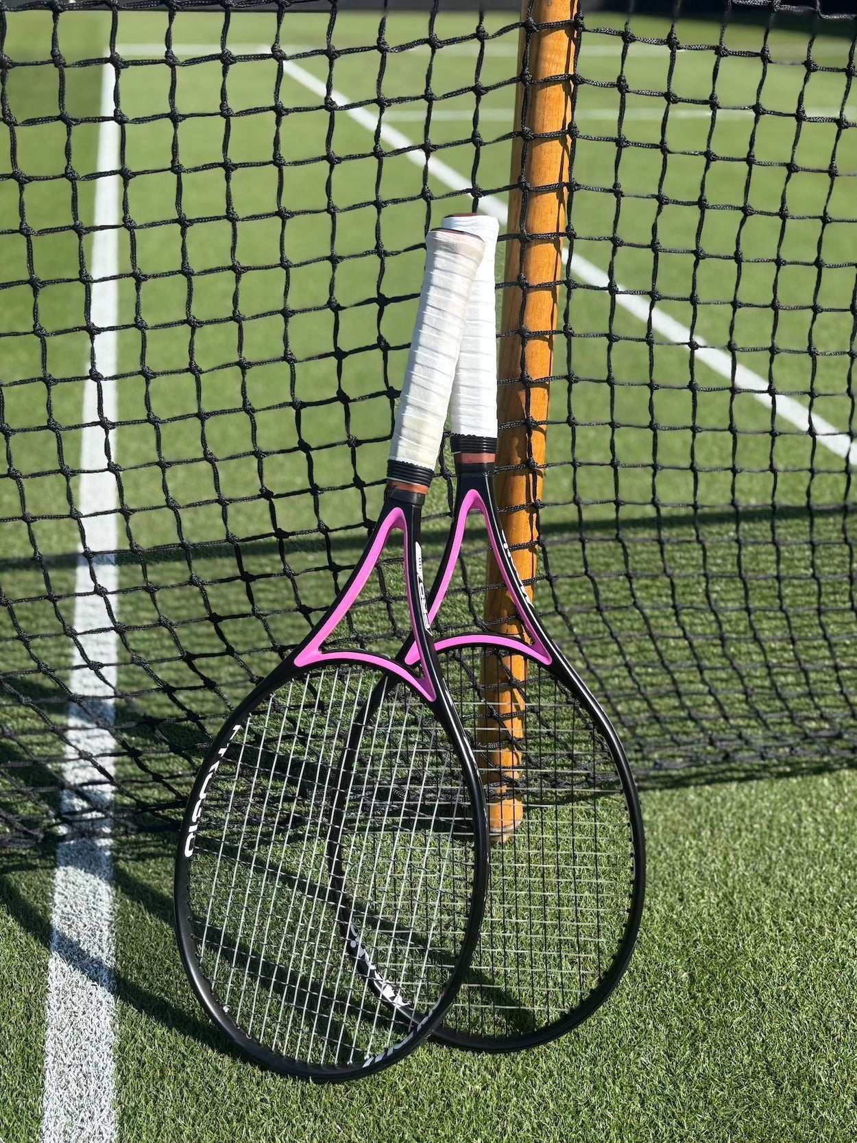 Two tennis racquets with white grips leaning against a net on a grass tennis court, illustrating tennis racquet stringing tension concepts.