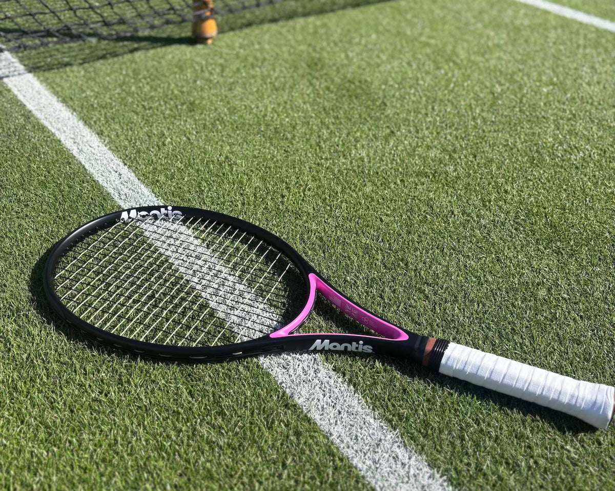 Mantis beginner tennis racket with black and pink frame lying on artificial turf near net line