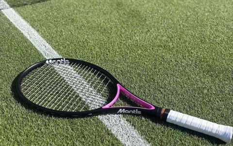 Mantis beginner tennis racket with black and pink frame and white grip lying on grass tennis court near net line