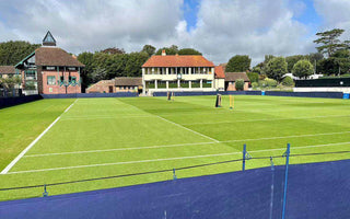 Outdoor tennis court with green grass, white court lines, and club buildings in the background on a sunny day