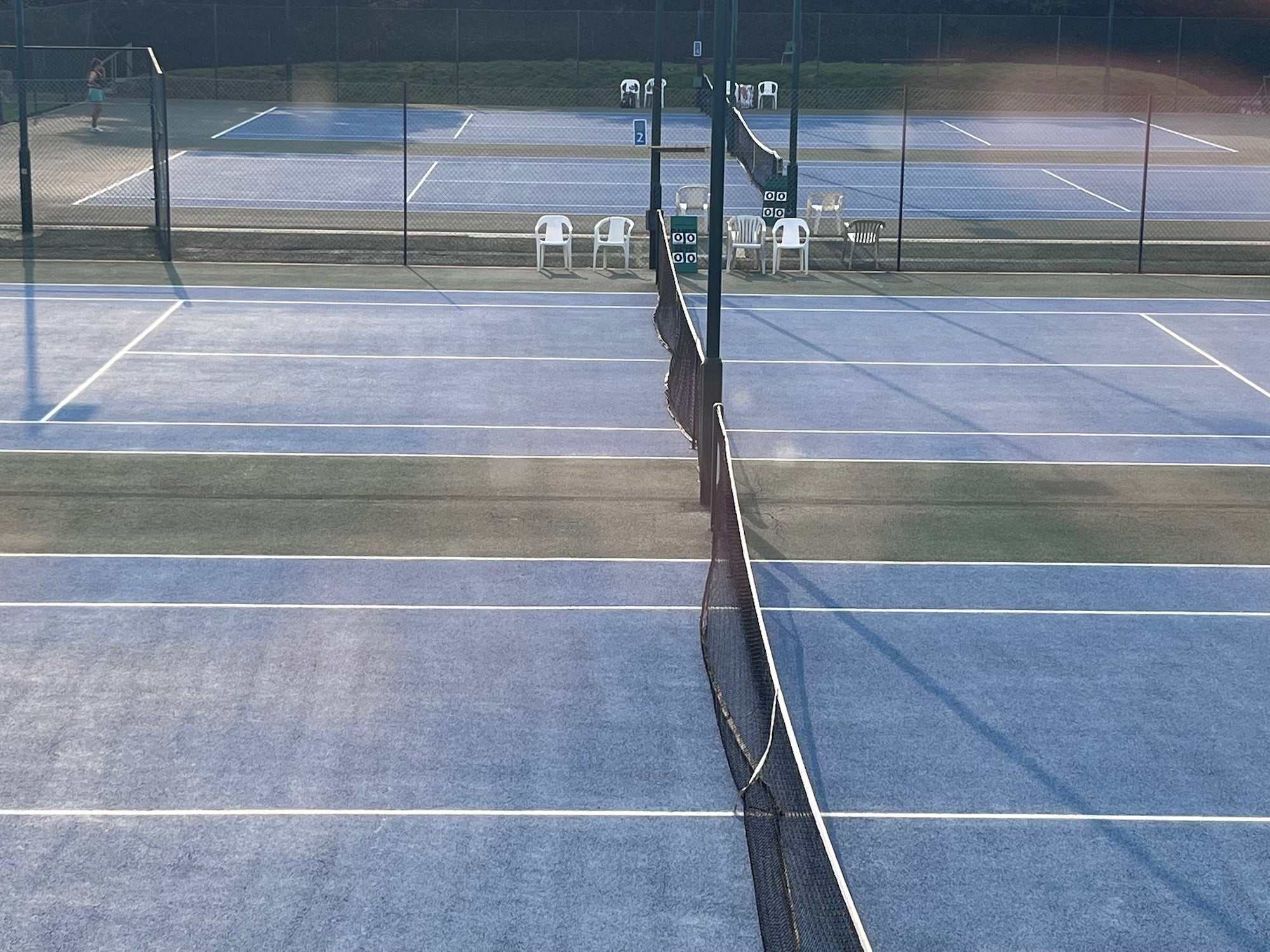 Outdoor blue hard tennis court surface with net and chairs visible at the far end