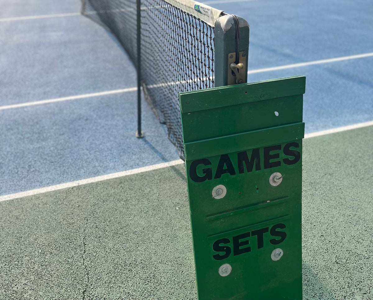 Green manual tennis scoreboard showing games and sets near net on blue court