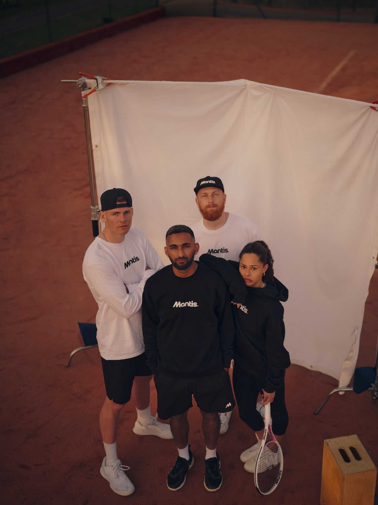 Group of diverse young adults wearing Mantis sportswear standing on a clay tennis court holding a tennis racket, promoting tennis for beginners.