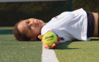 Close-up of a person lying on a hard tennis court holding a Mantis hard court tennis ball along the court's white line