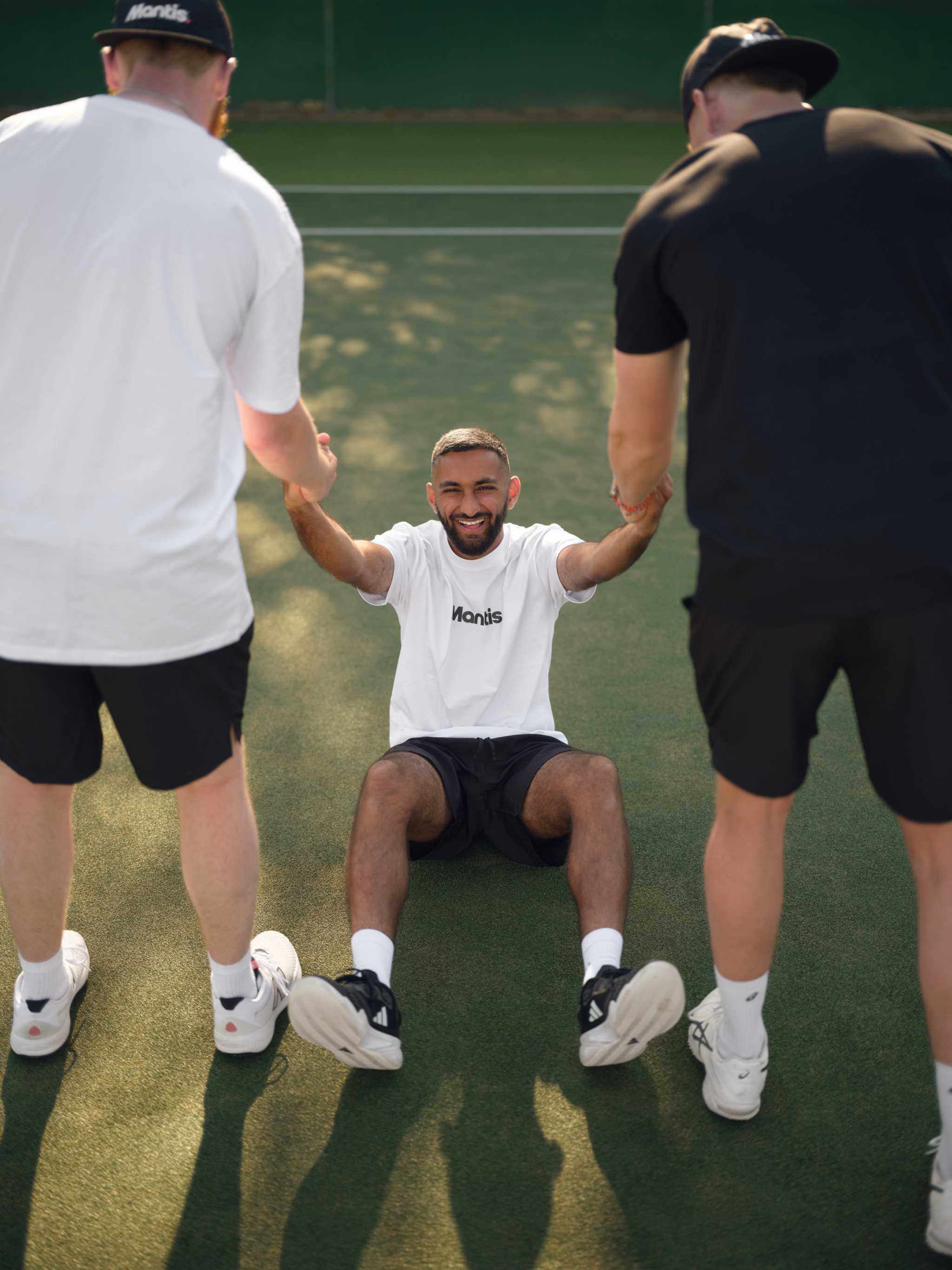 Three men on tennis court performing strength training exercise with one man sitting and two men helping him stand up