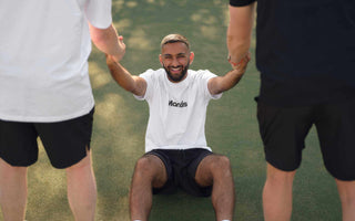 Three tennis players performing strength training exercises on court, one sitting and being assisted by two standing teammates during a workout session