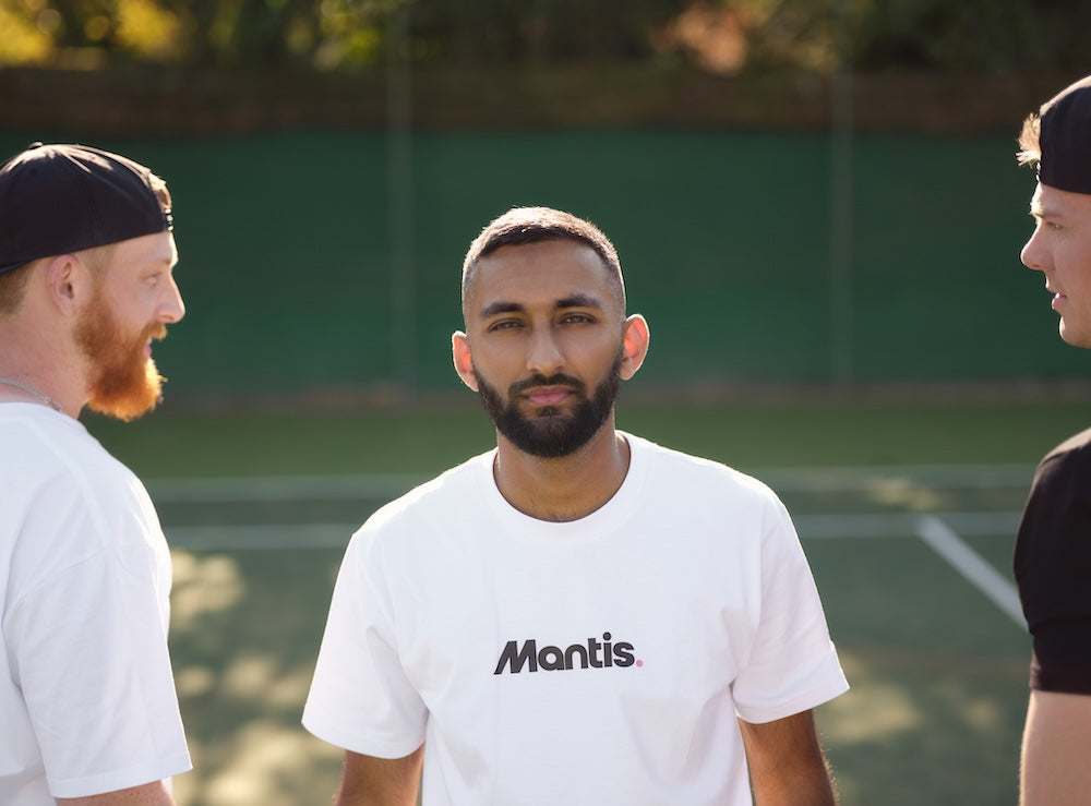 three men discussing on outdoor tennis court wearing casual sportswear with man in center wearing white Mantis t-shirt a modern guide to coaching tennis