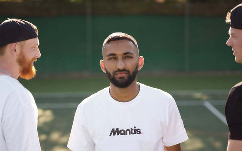 three men discussing on outdoor tennis court wearing casual sportswear with man in center wearing white Mantis t-shirt a modern guide to coaching tennis