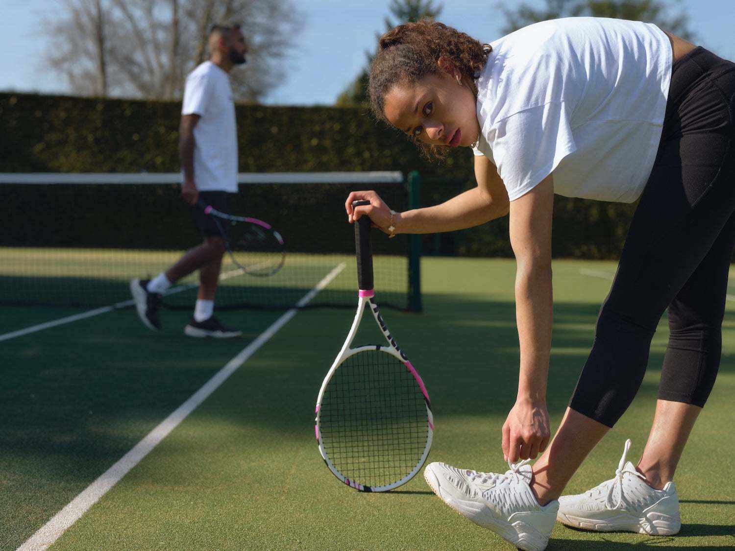 woman tying shoelaces with tennis racket on tennis court preparing for game with male player in background