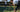 woman tying shoelaces on tennis court holding racket with man in background practicing tennis