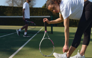 Woman tying shoelace on tennis court holding racket, man with racket in background practicing tennis