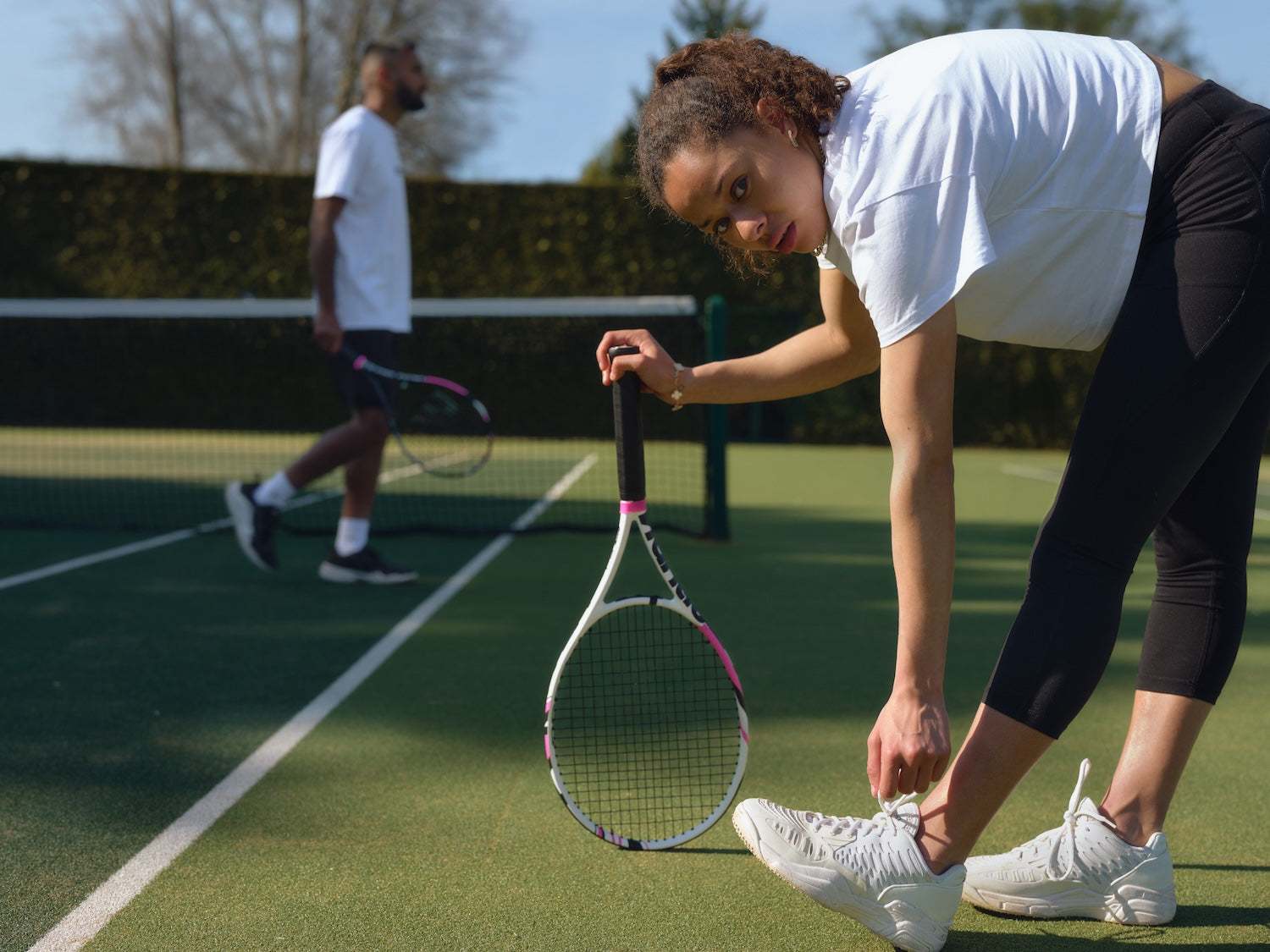 Woman tying shoelaces on tennis court holding a racket, man practicing in the background during tennis training session