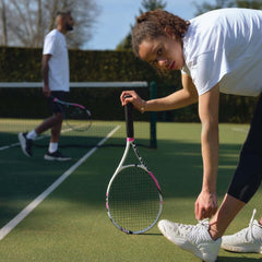 Woman tying shoelaces on tennis court holding a racket, man practicing in the background during tennis training session