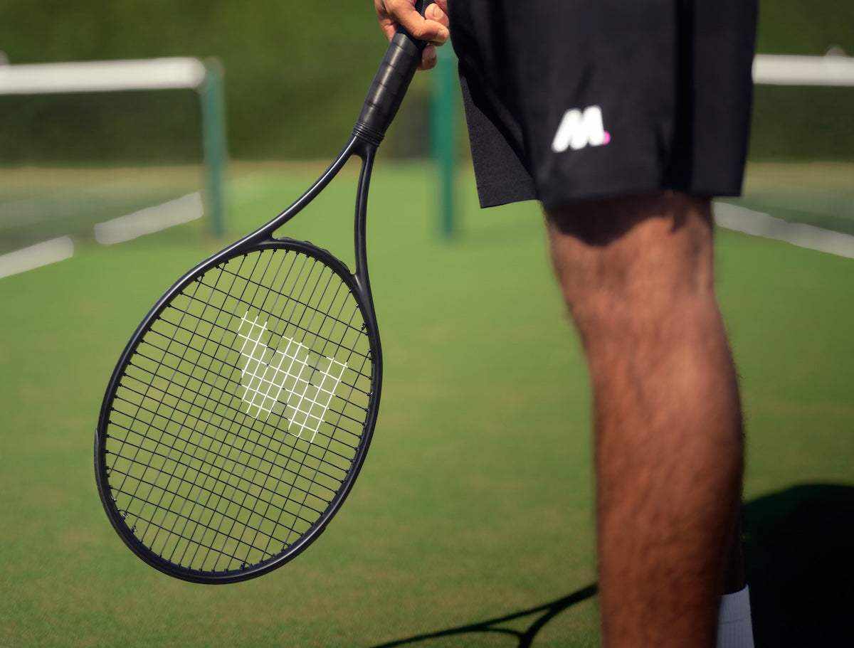 Close-up of a person holding a black tennis racket on an outdoor tennis court, focusing on the racket strings and grip, illustrating restring a tennis racket.