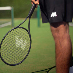Close-up of a person holding a black tennis racket on an outdoor tennis court, focusing on the racket strings and grip, illustrating restring a tennis racket.