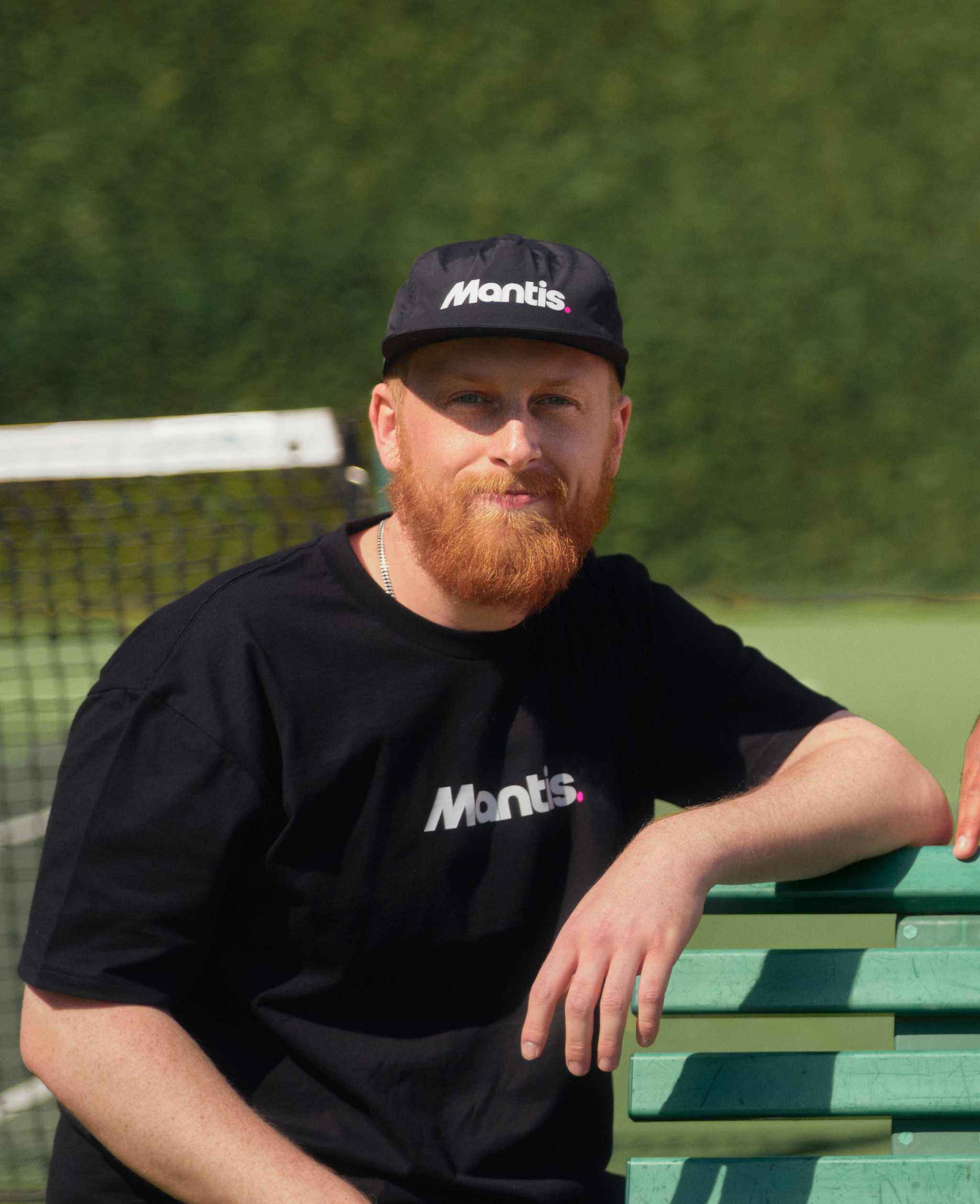 Man wearing black Mantis Snapback Cap and matching Mantis t-shirt sitting on bench at tennis court