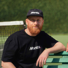 Man wearing black Mantis Snapback Cap and matching Mantis t-shirt sitting on bench at tennis court