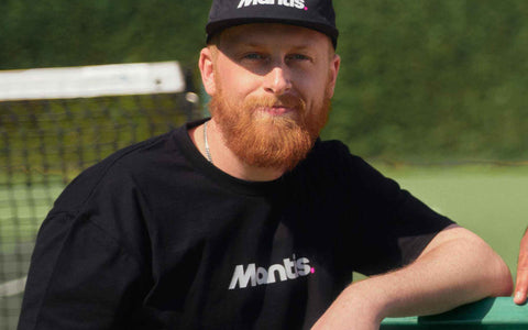 Man wearing black Mantis Snapback Cap and matching Mantis t-shirt sitting on bench at tennis court