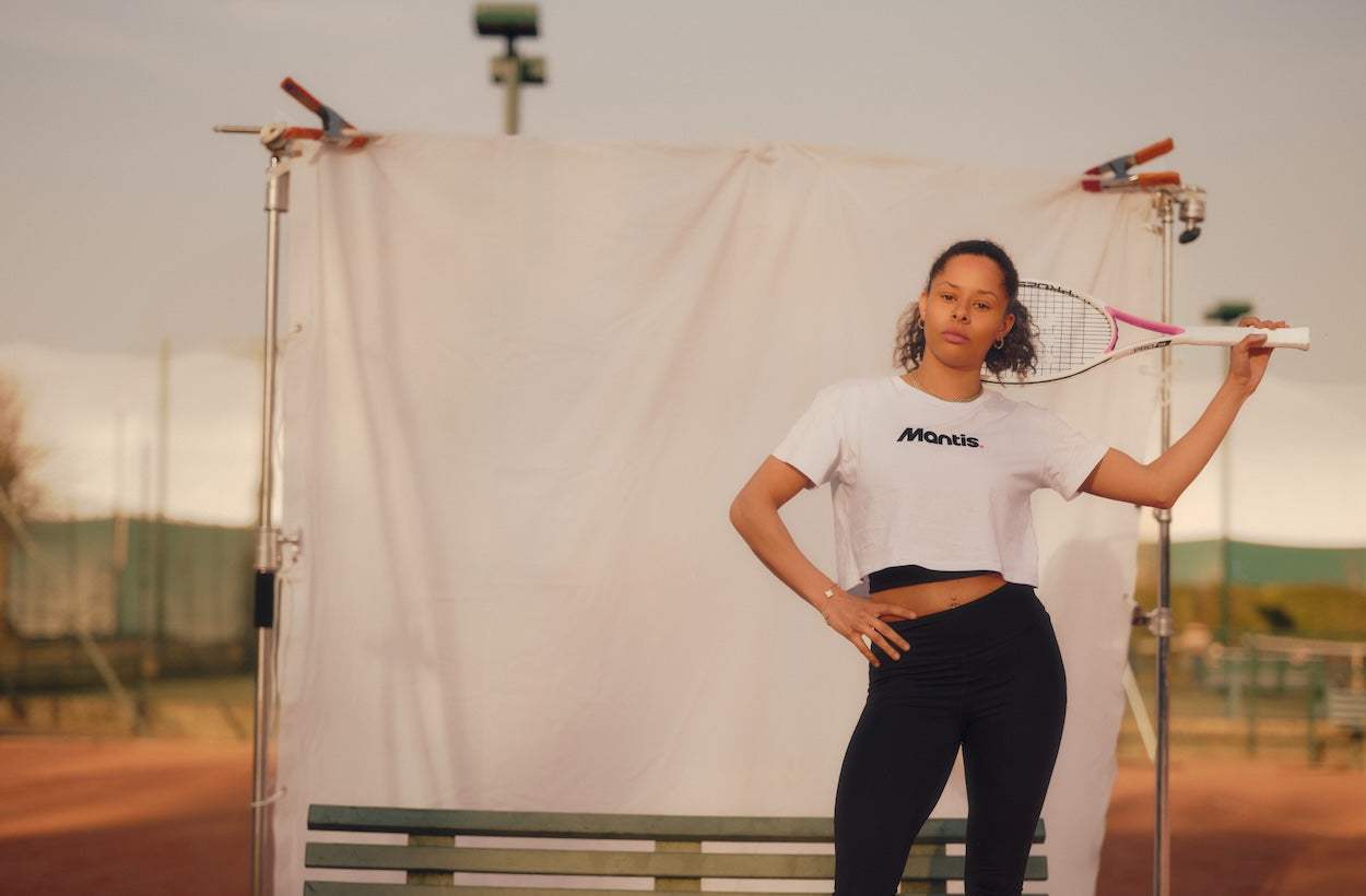 woman holding tennis racket on shoulder outdoors wearing mantis shirt learning how to play tennis guide