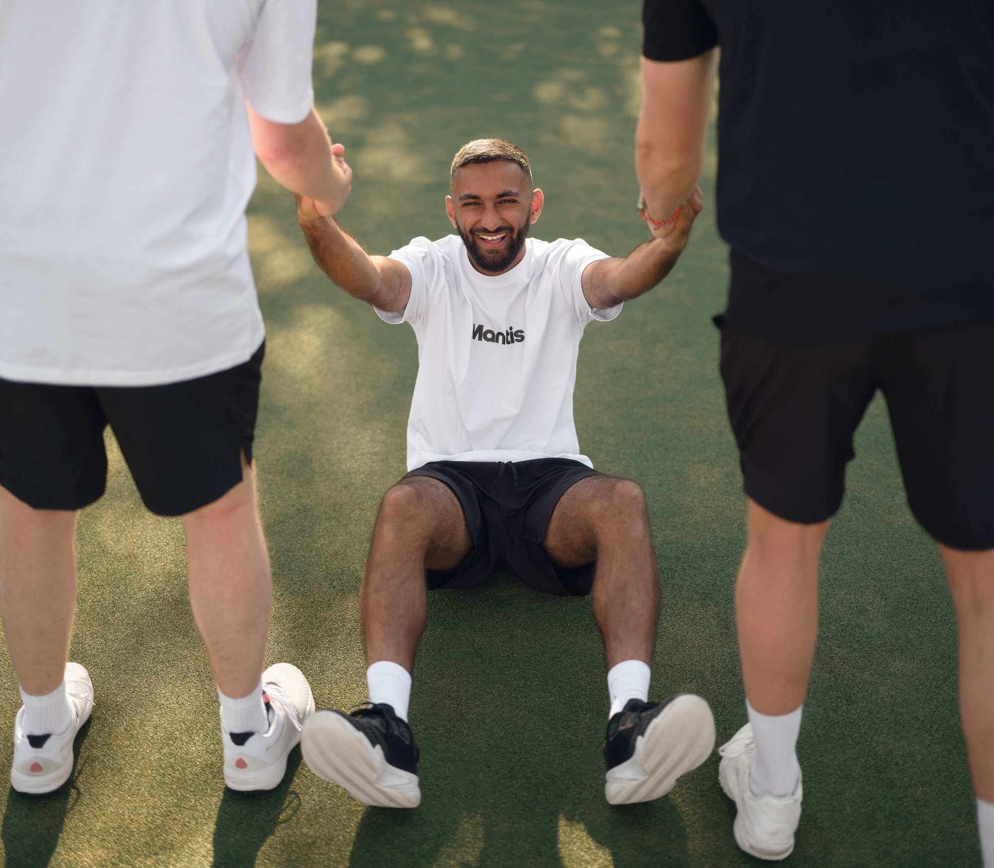 Young man sitting on tennis court holding hands with two standing people helping him up Wimbledon tickets guide and tennis experience