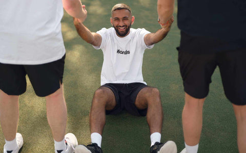 Young man in white Mantis tennis shirt sitting on court holding hands of two standing people in black and white attire