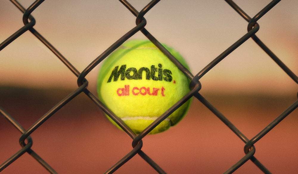Close-up of a yellow Mantis all court tennis ball caught in a chain-link fence at sunset