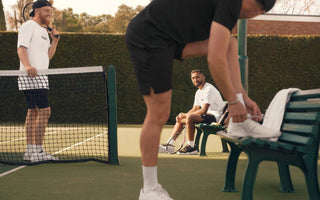 Three men on a tennis court preparing for a game, one tying shoes on a bench with tennis rackets nearby, illustrating online tennis lessons and coaching.