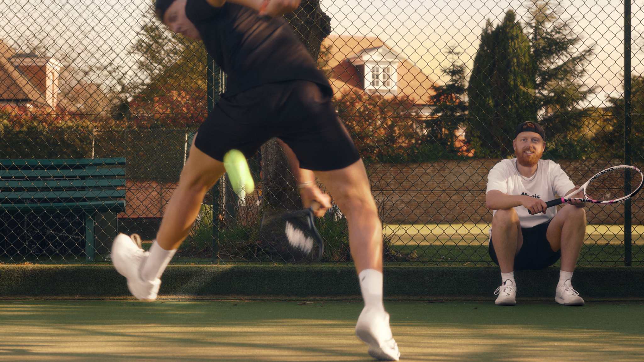 Tennis player performing a dynamic warm-up on court with another player sitting nearby holding a racket, practicing essential tennis warm up exercises