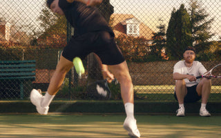 Two tennis players warming up on a court, one in mid-swing hitting a tennis ball and the other sitting with a racket, demonstrating tennis warm up exercises.