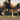 Tennis player executing a backhand shot on an outdoor court with a coach watching in the background