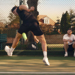 Tennis player executing a backhand shot on an outdoor court with a coach watching in the background