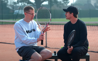 Two people discussing tennis rackets on a clay tennis court.