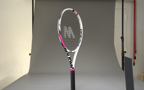 White and pink tennis racket standing upright in a professional studio setting against a gray backdrop.